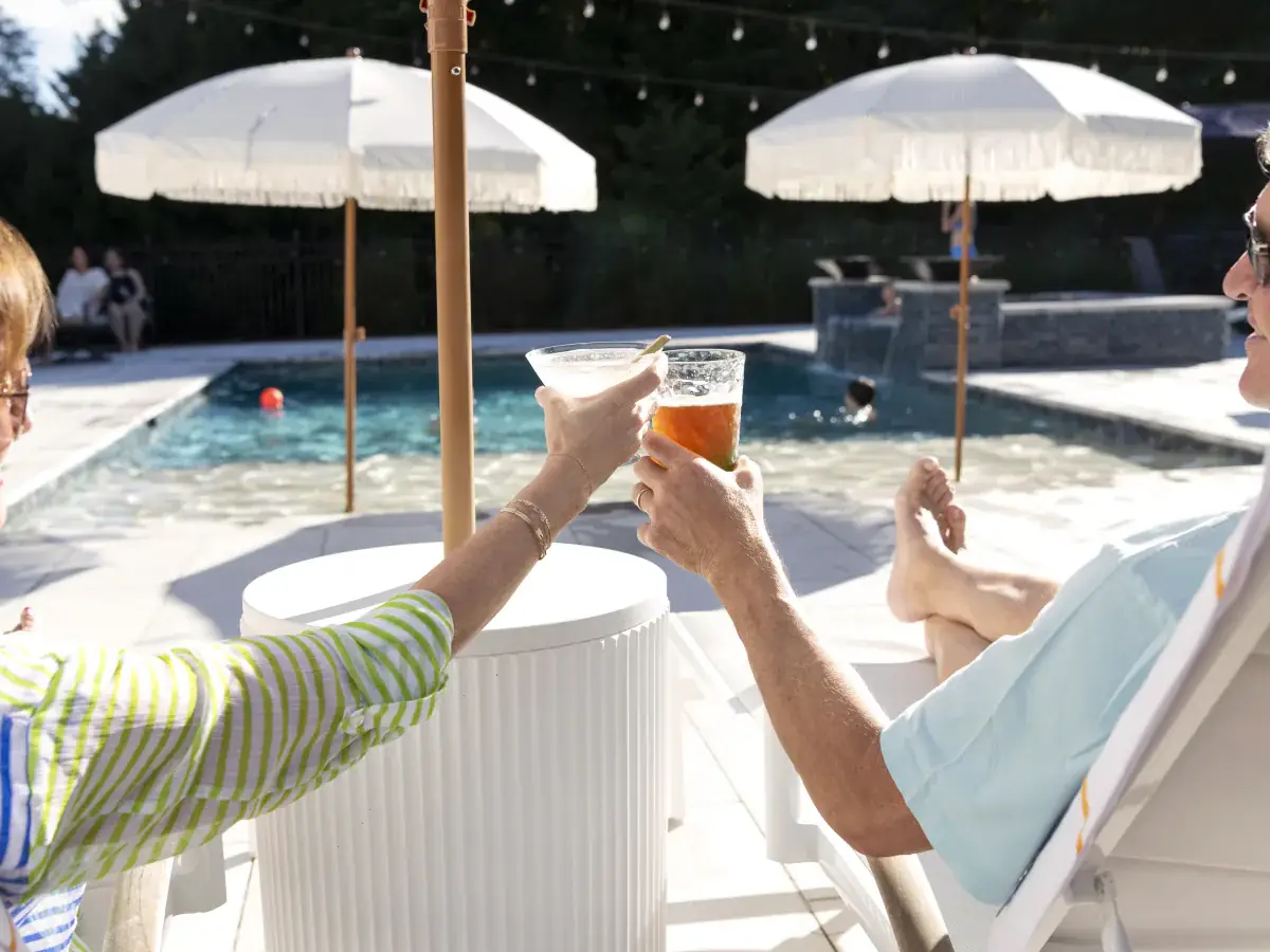 A couple toasting drinks in lounge chairs beside their RutherfordMade pool under fringed umbrellas